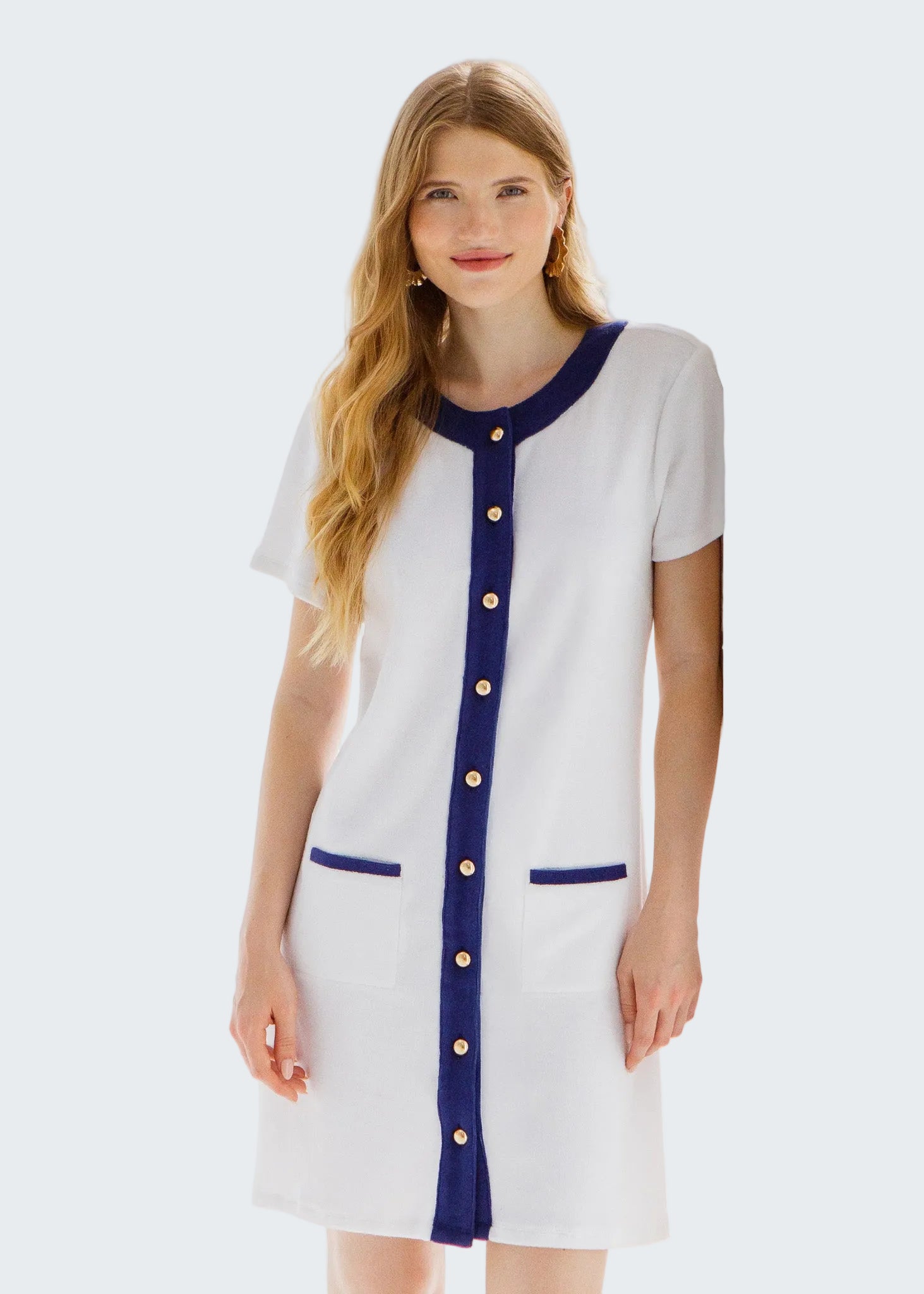 Woman wearing a white dress with blue accents and gold buttons in a bright room.