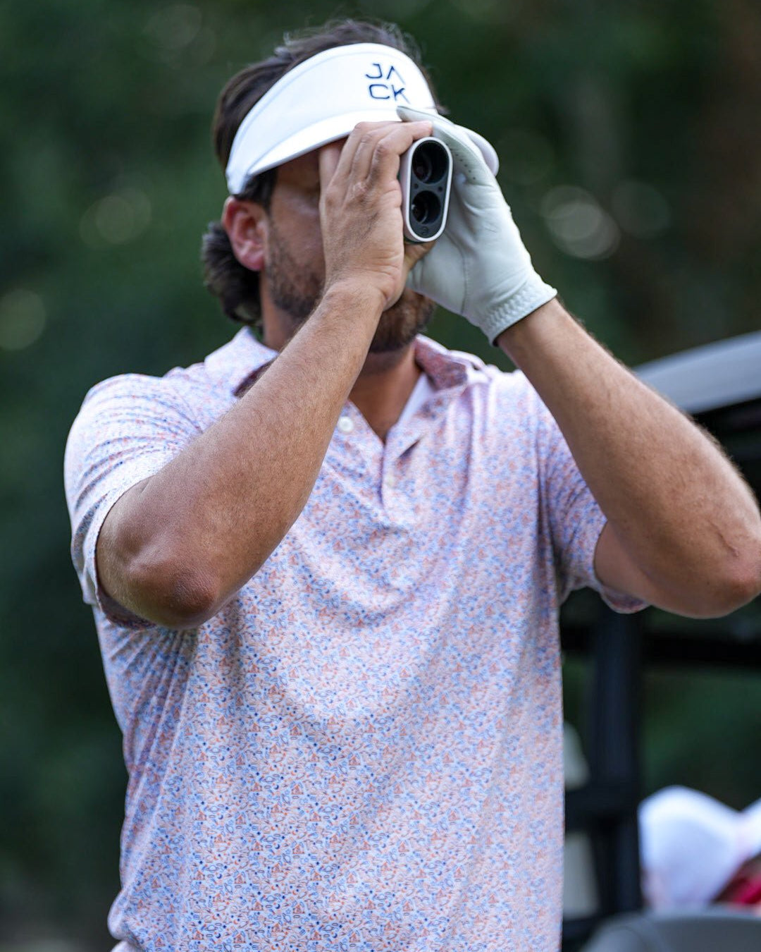 Man in a patterned shirt and visor holding binoculars with a blurred natural background
