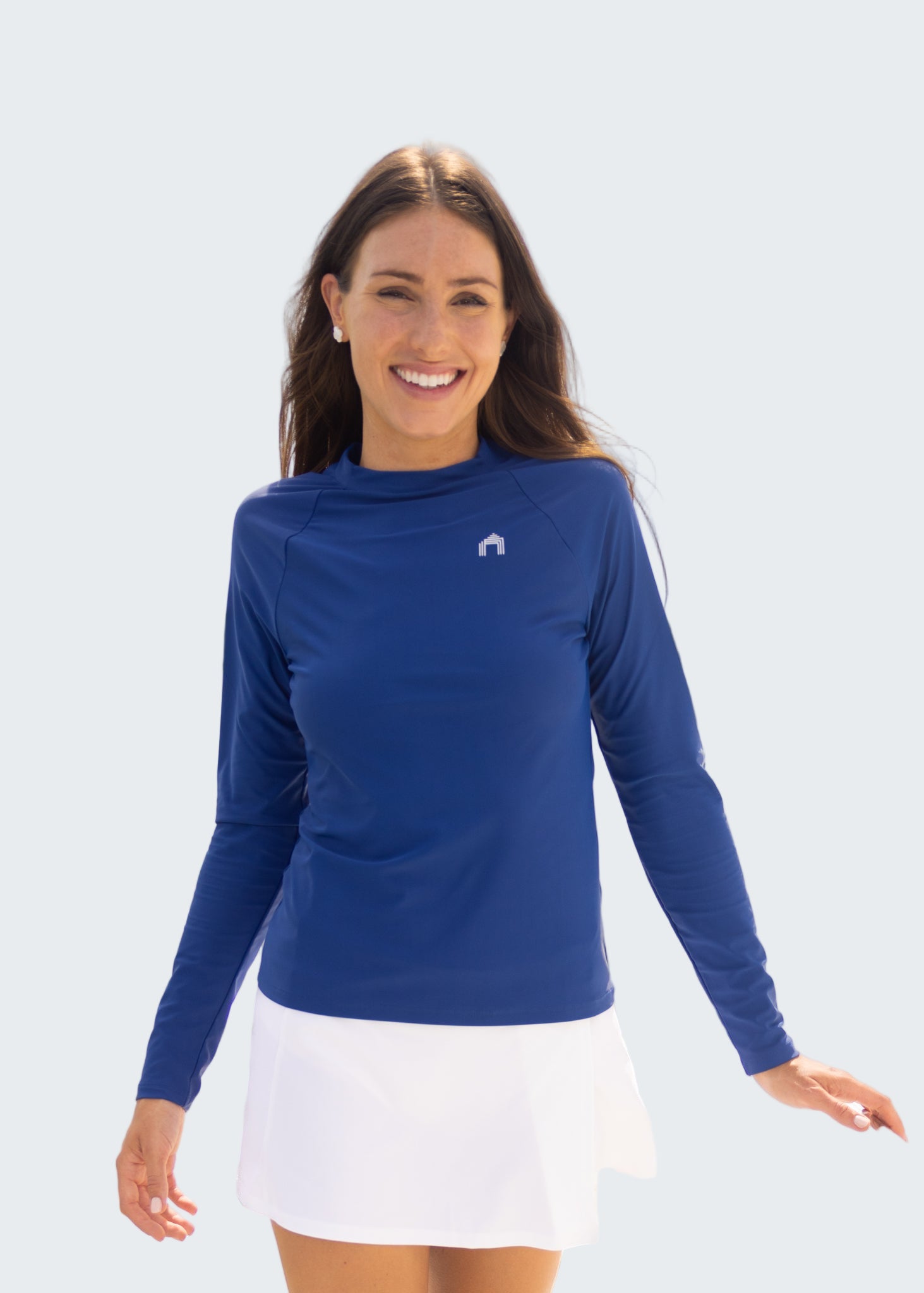 Woman wearing a blue long-sleeve shirt and white skirt standing by a body of water with sailboats in the background.