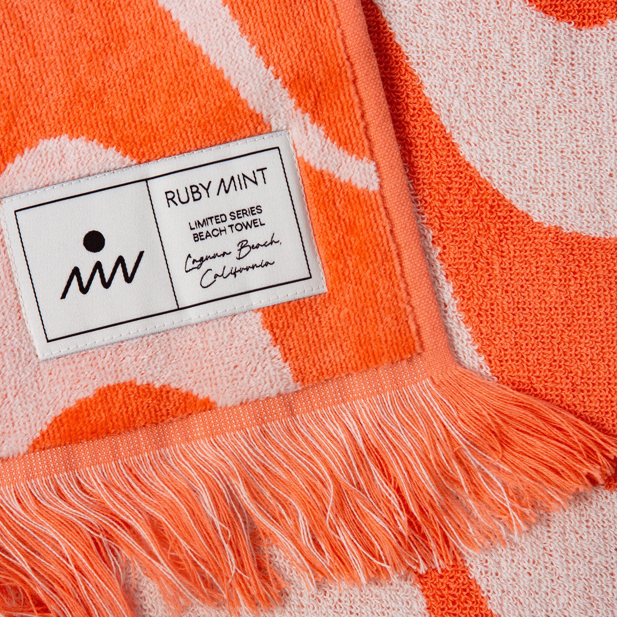 Close-up of a Ruby Mint beach towel with a label on an orange and white patterned background.
