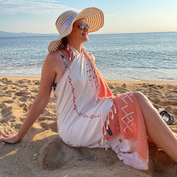 Woman sitting on a beach with a towel draped over her, wearing a hat and sunglasses.