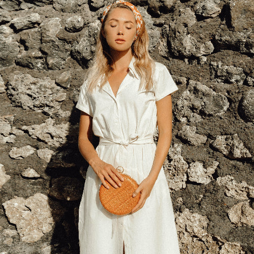 Woman in a white dress holding an orange clutch against a stone wall.