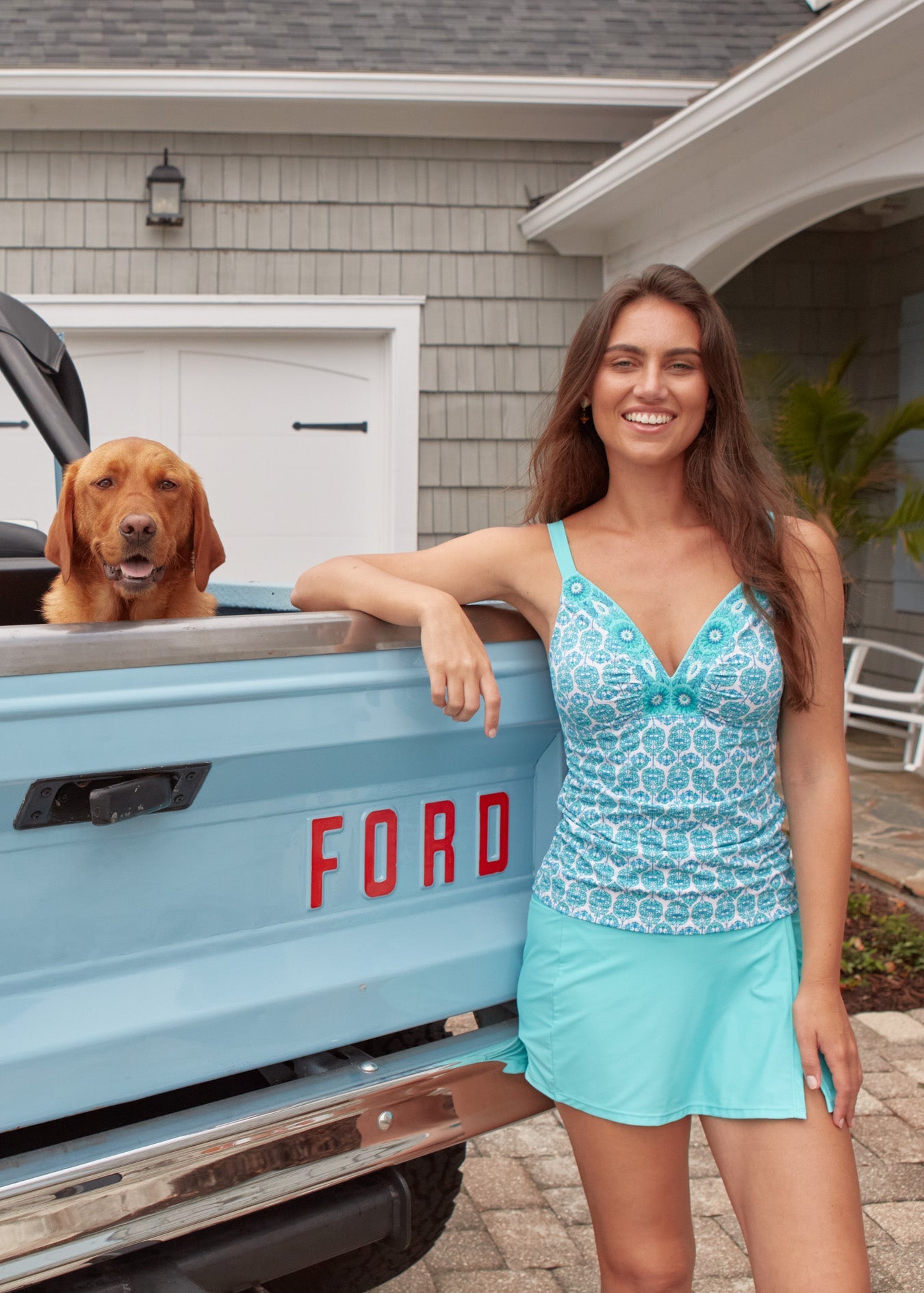 Woman in a blue dress standing next to a Ford truck with a dog inside