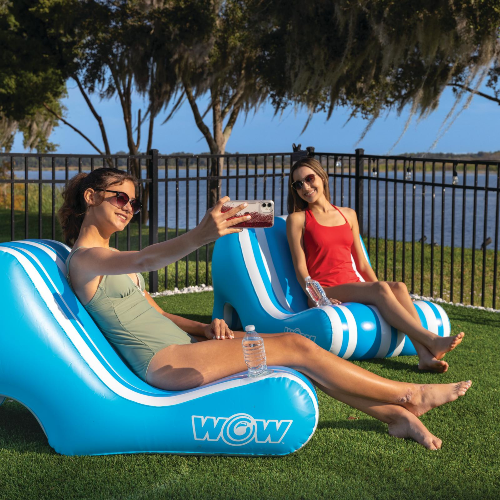 Two women sitting on inflatable lounge chairs by a lake, taking a selfie.