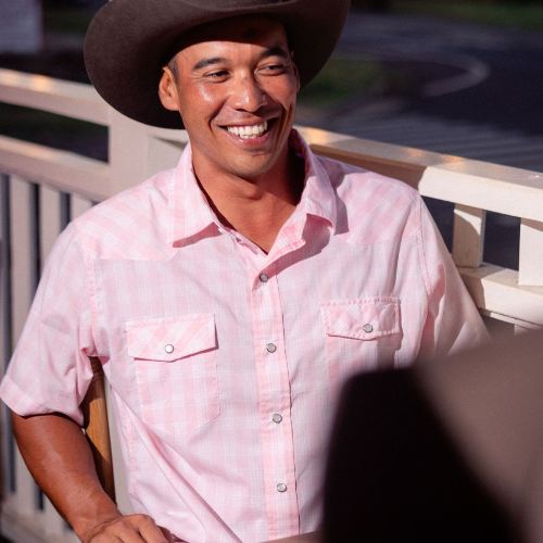 Man wearing a pink shirt and cowboy hat smiling outdoors.