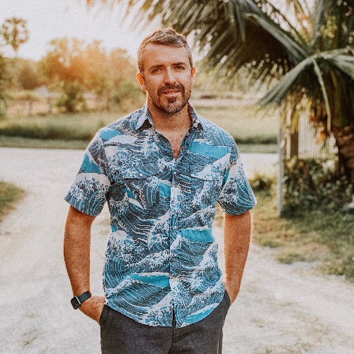 Man wearing a blue patterned shirt standing outdoors with palm trees in the background