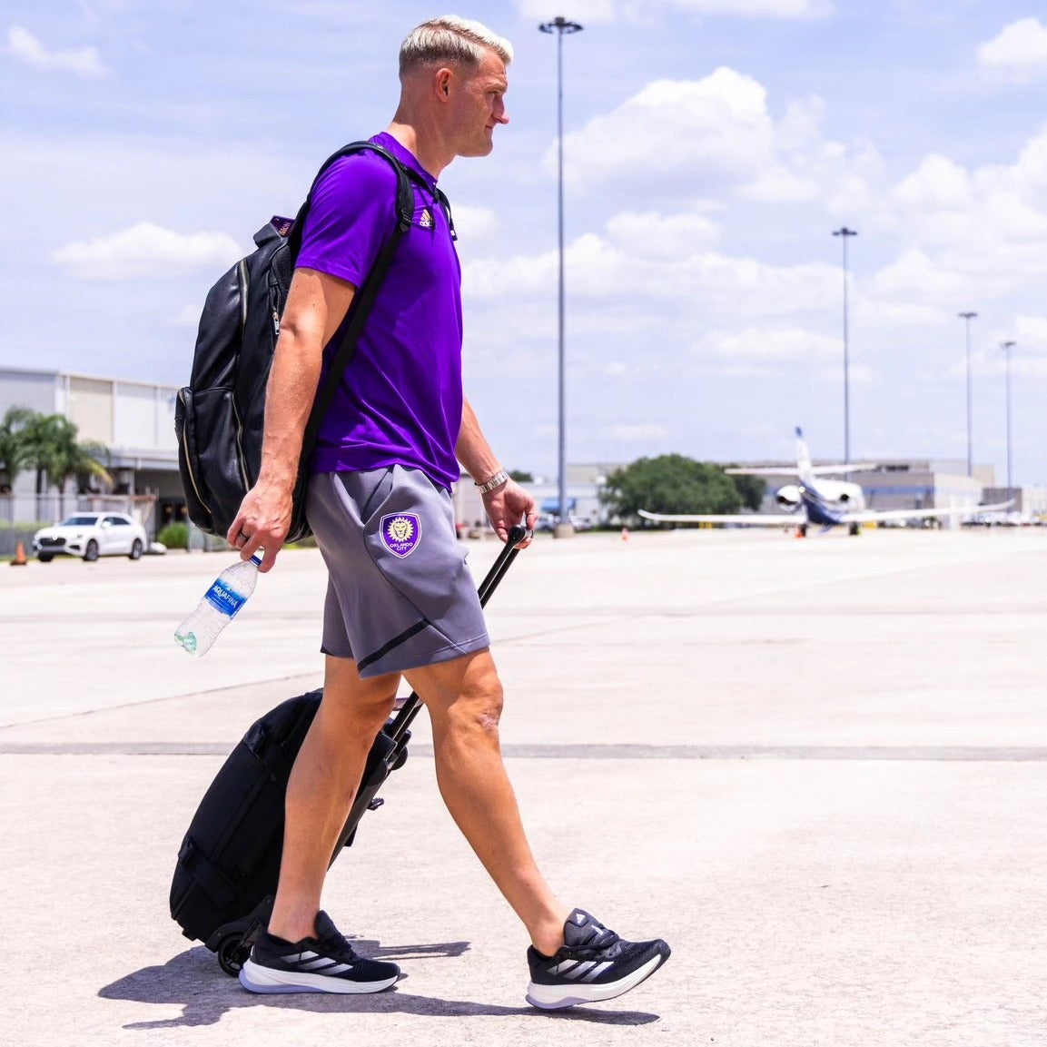 Person in purple shirt and gray shorts walking with a suitcase at an airport.