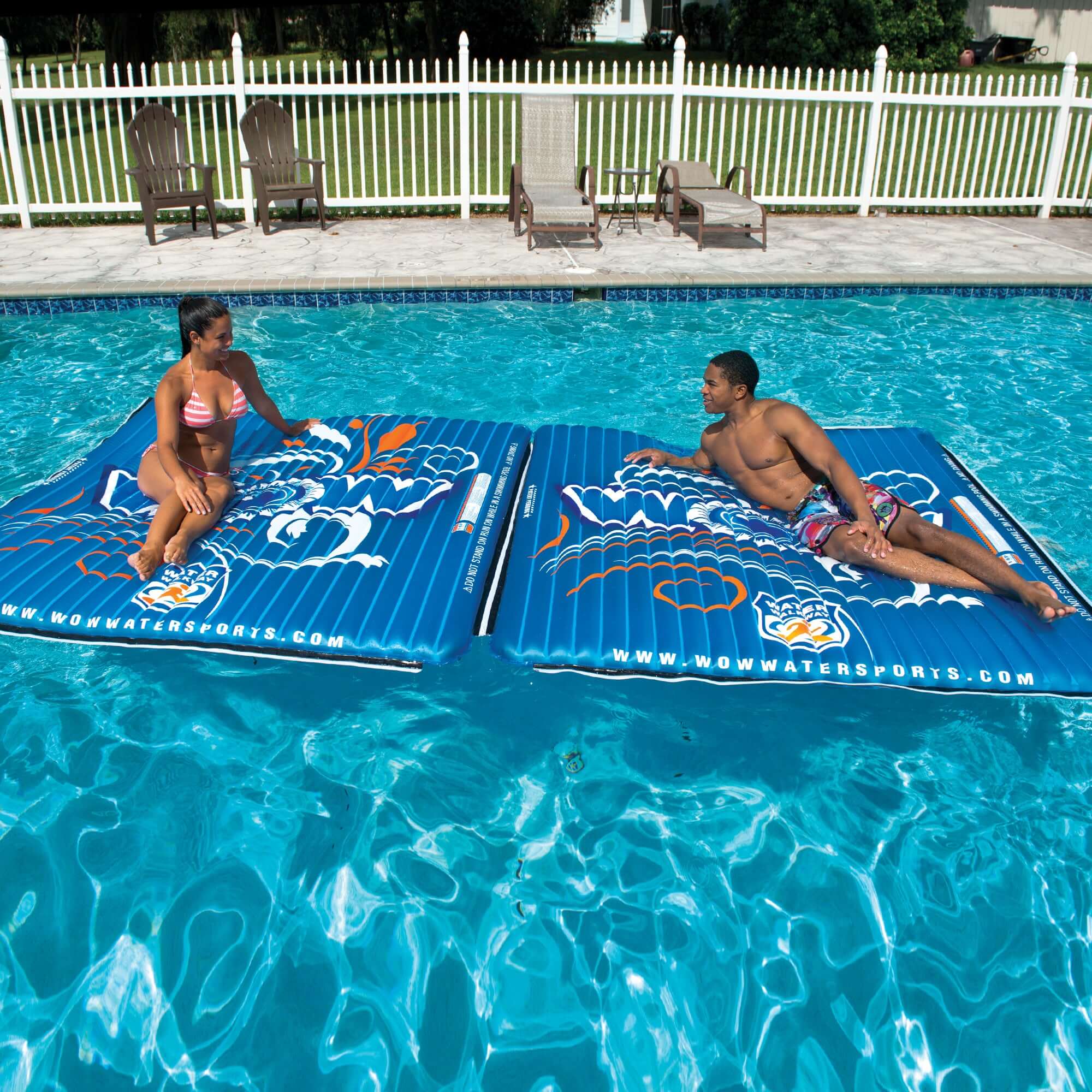 Two people on inflatable pool floats in a swimming pool with a white fence in the background.