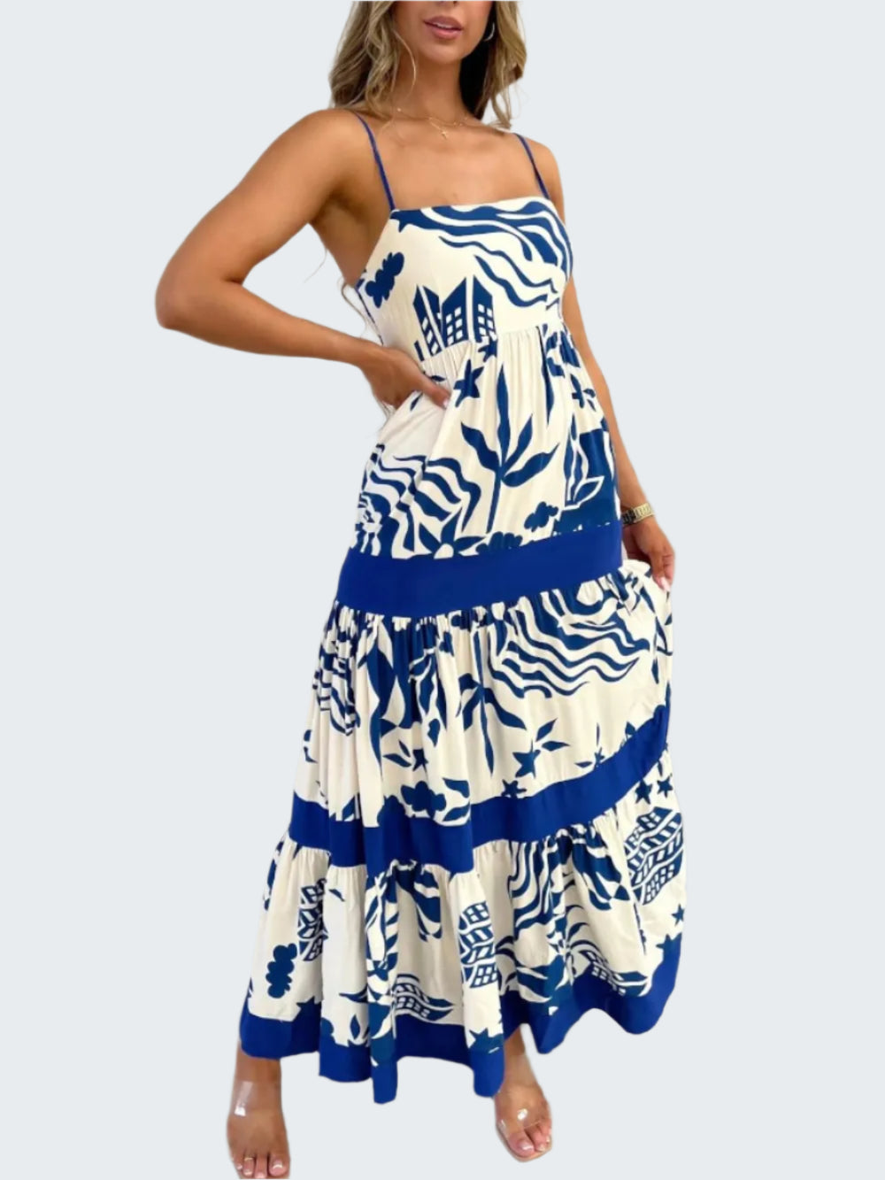 A model wearing a square neck, floral printed maxi dress with a blue belt and trim, standing in a room with a white background.