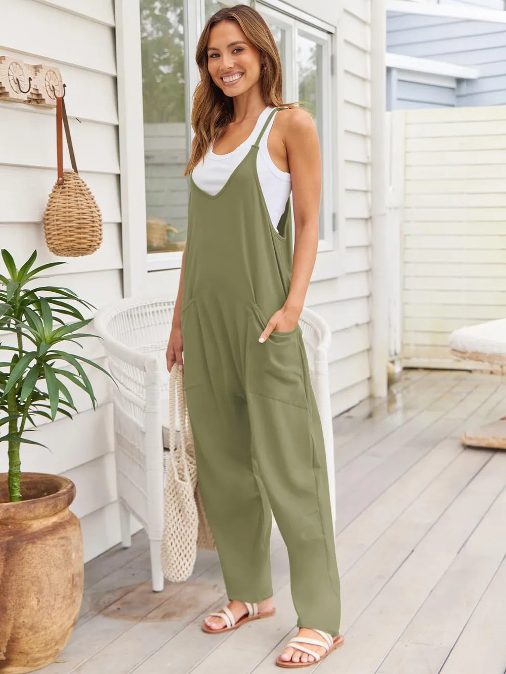A woman standing on a patio wearing a black sleeveless jumpsuit with a white tank top underneath, holding a wicker handbag and wearing white sandals.
