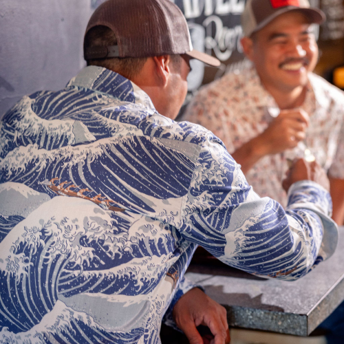 Two men sitting at a table with one wearing a blue wave-patterned shirt.