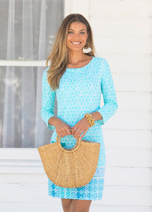 Woman wearing a blue patterned dress holding a straw bag against a white background