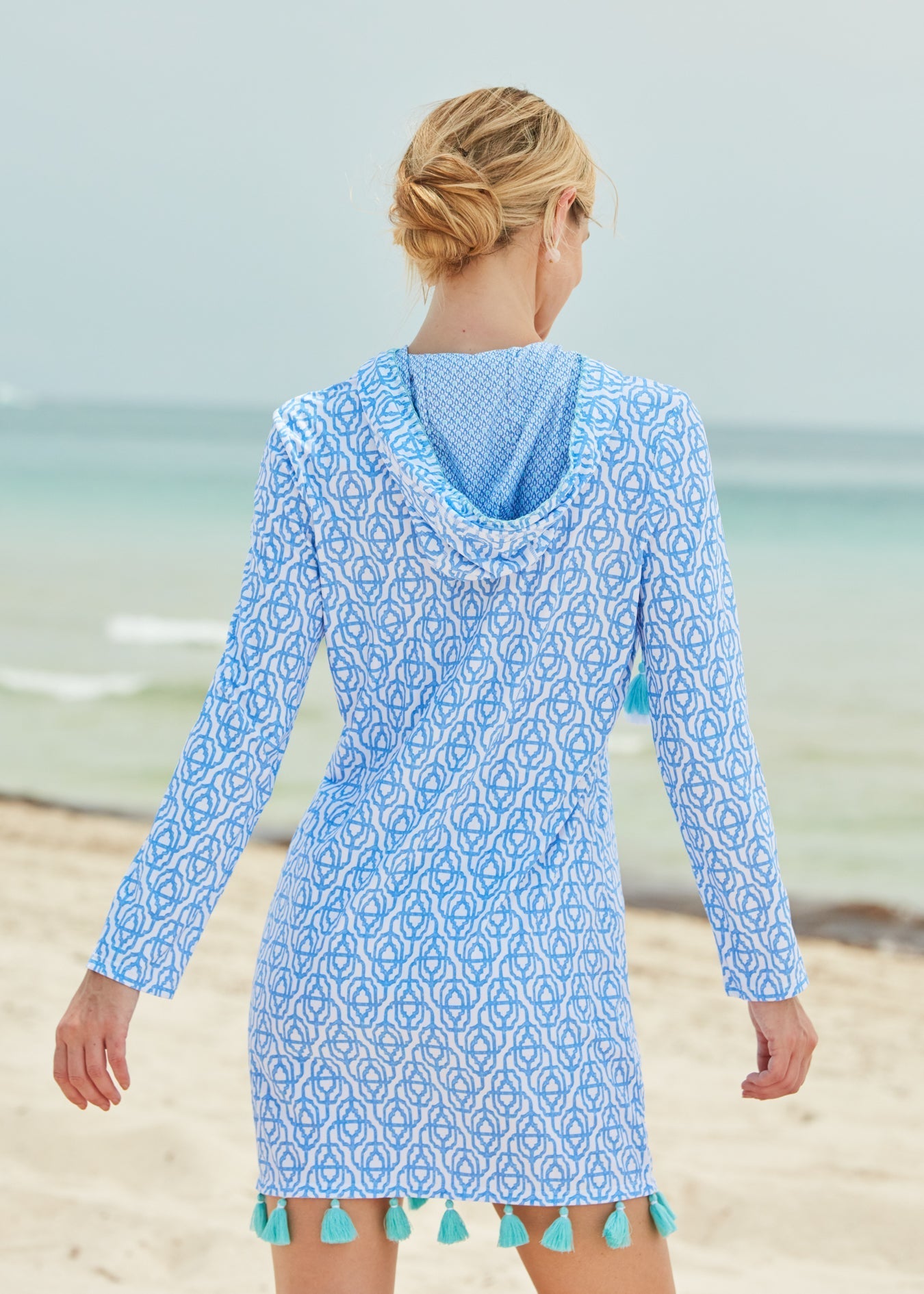 Woman wearing a blue patterned dress with hood and green pom-poms on a beach.