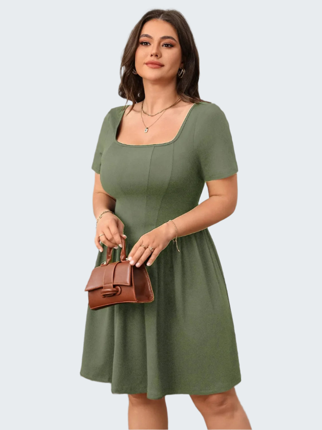 A model wearing a green square neck, short sleeve, mini dress with a waistband, standing in a room with a neutral decor.