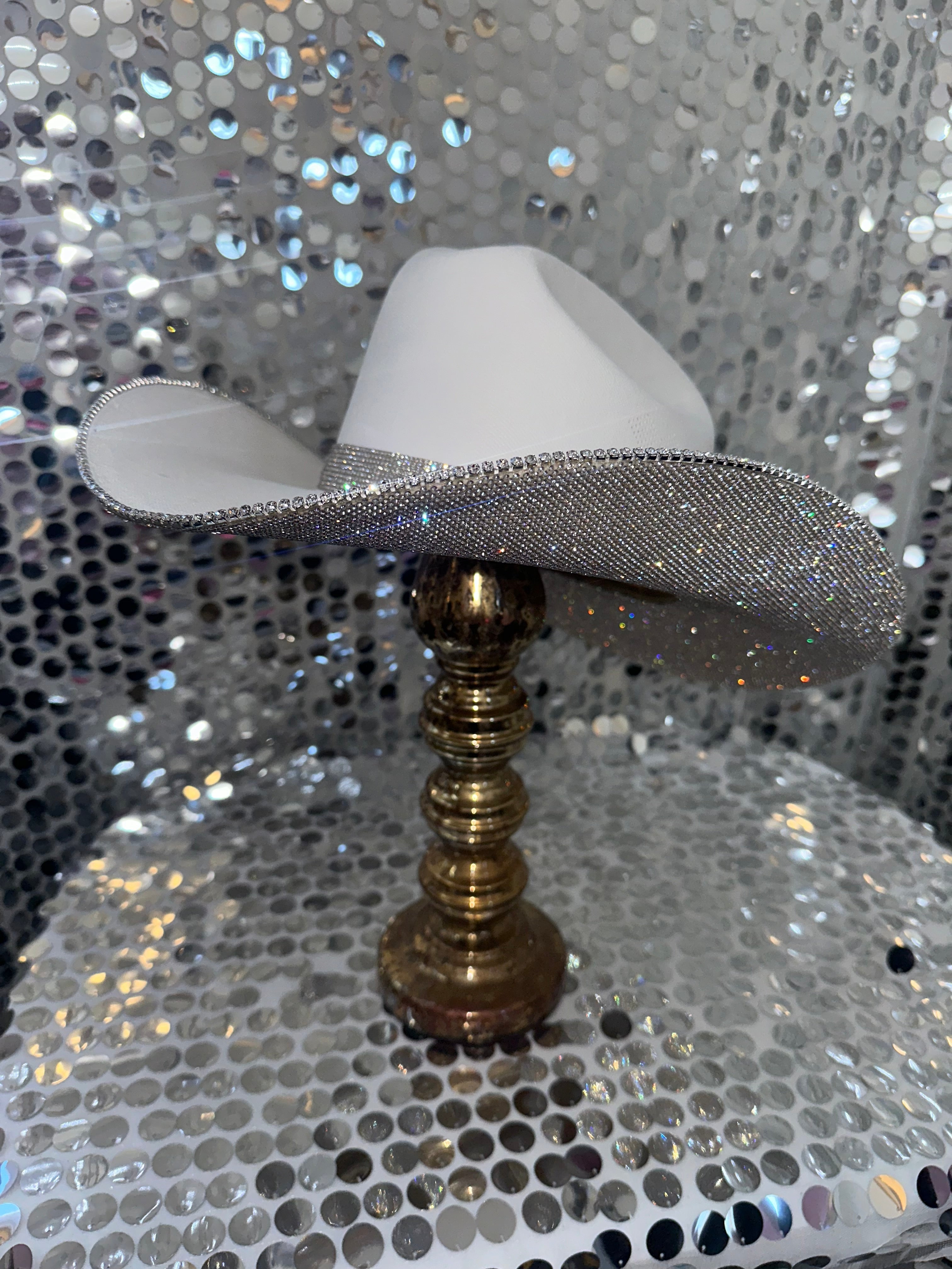 White cowboy hat with a silver brim on a gold stand against a silver sequin background