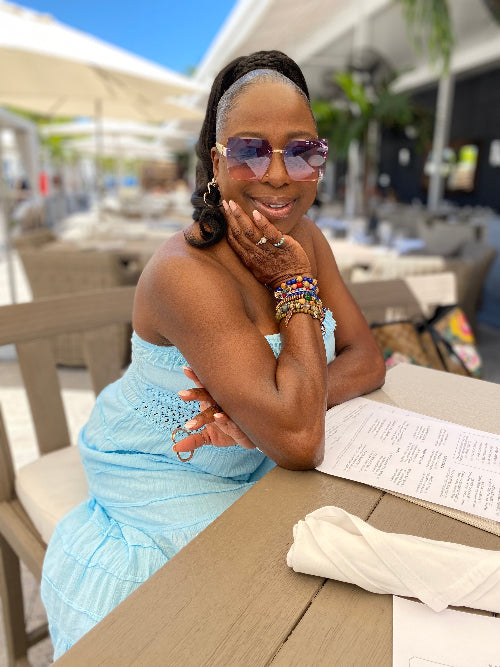 Woman in a light blue dress and sunglasses sitting at an outdoor table with a menu.