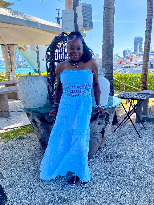Woman in a blue dress standing outdoors with palm trees and a city skyline in the background.