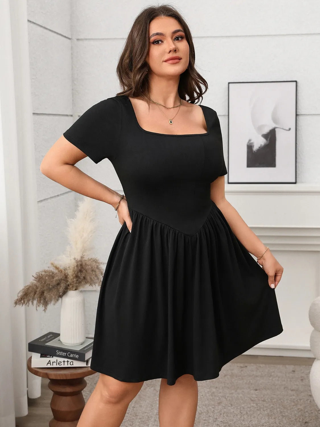 A model wearing a green square neck, short sleeve, mini dress with a waistband, standing in a room with a neutral decor.
