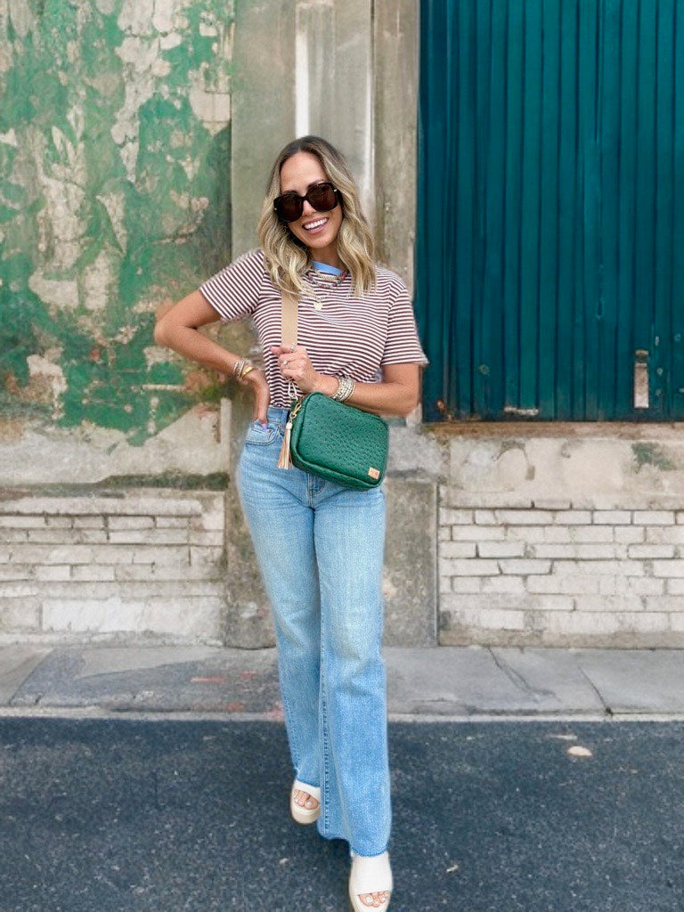 Woman in striped shirt and jeans holding a green handbag against a textured wall.