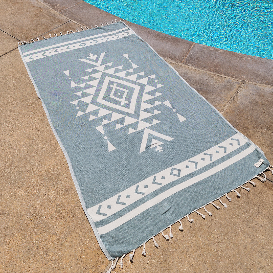 Blue and white patterned towel with tassels on a stone surface near a pool.