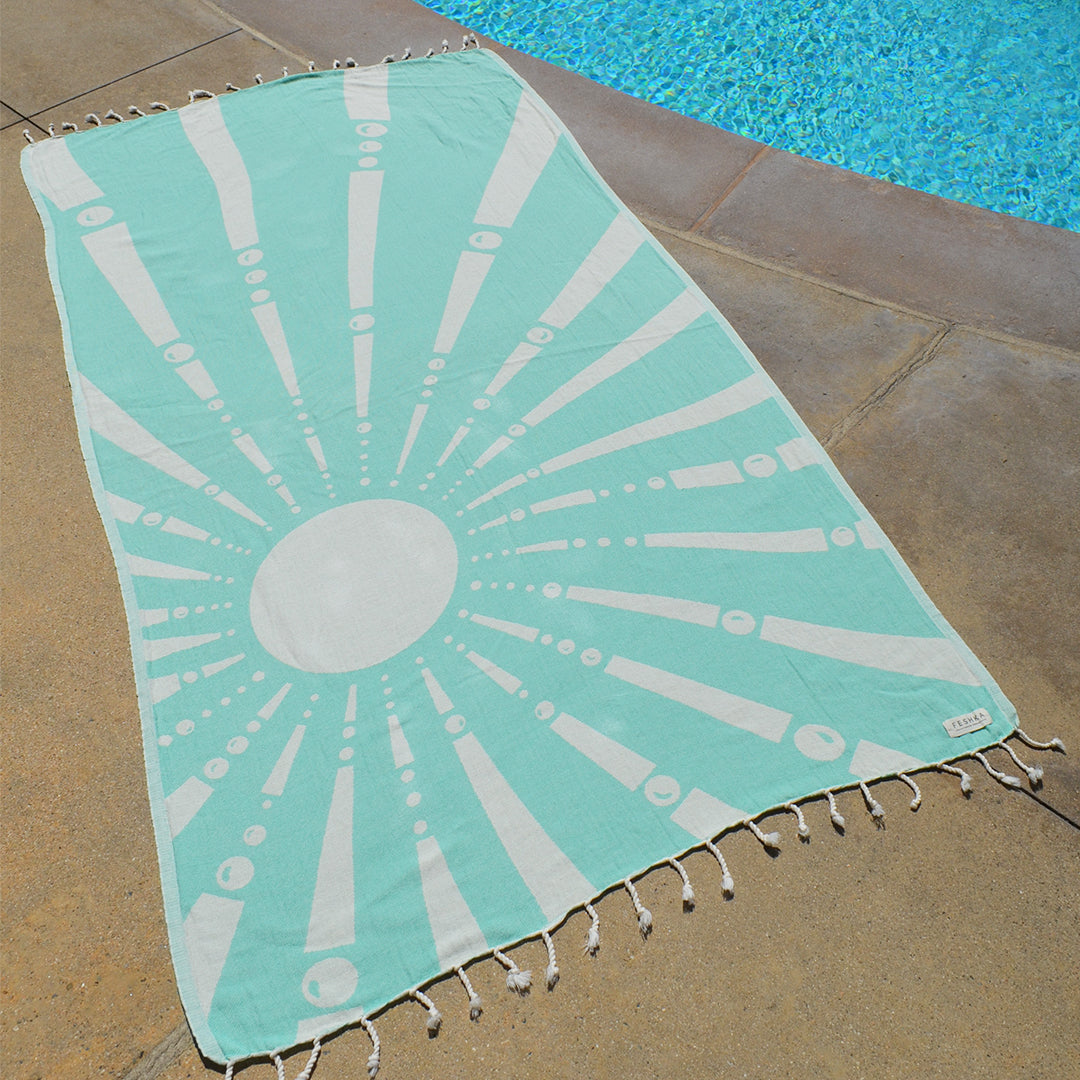 Turquoise and white beach towel with sunburst pattern on a stone surface near a pool.