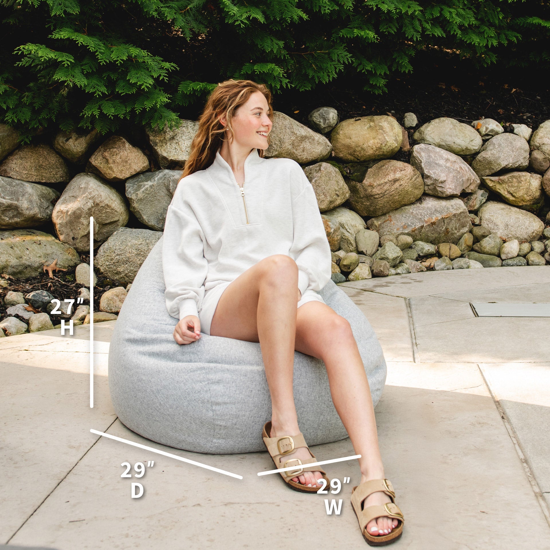 Woman sitting on a gray bean bag chair with measurements indicated, outdoors.