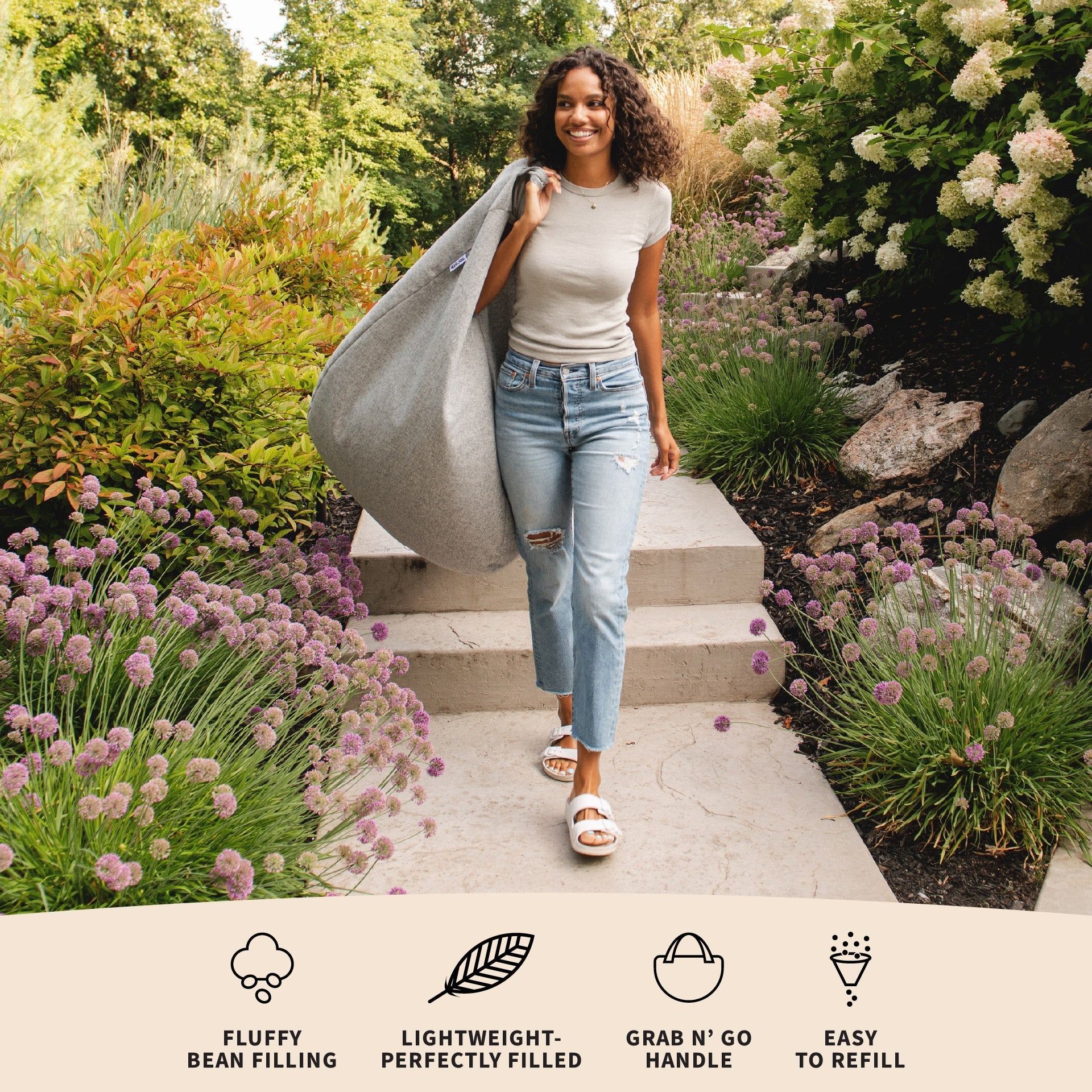 Woman walking outdoors with a large gray bag, surrounded by greenery.