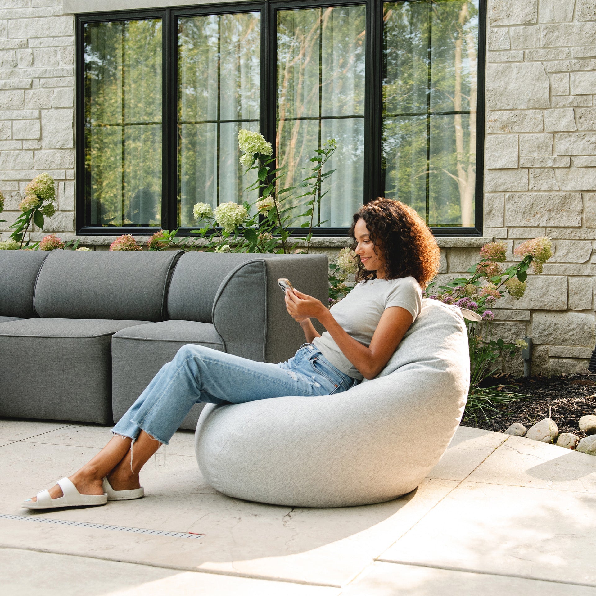 Woman sitting on a bean bag chair in an outdoor setting with a gray sectional sofa and stone wall.