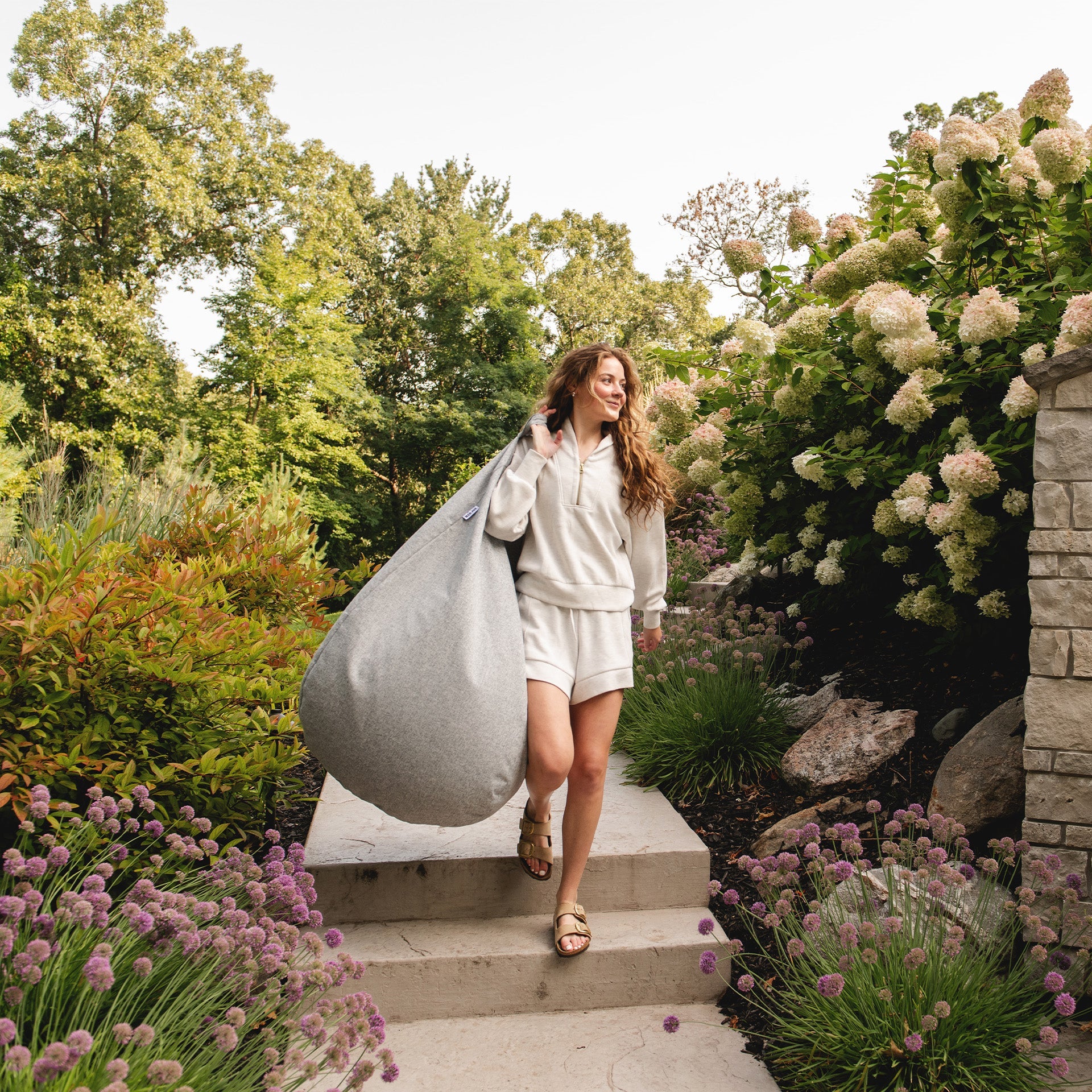 Woman carrying a large gray bean bag in a garden setting with flowers and trees.