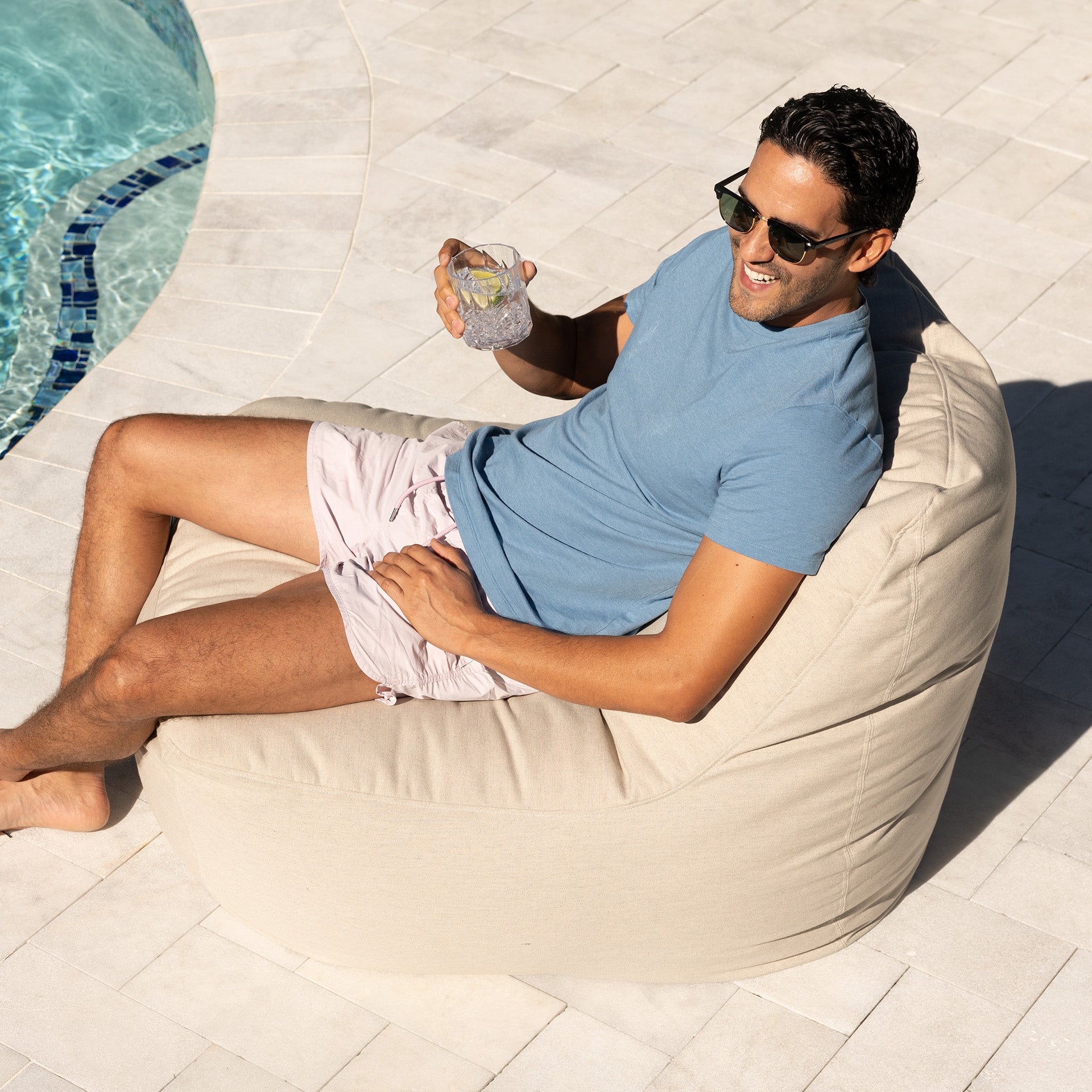 Man relaxing on a bean bag by a pool, holding a drink
