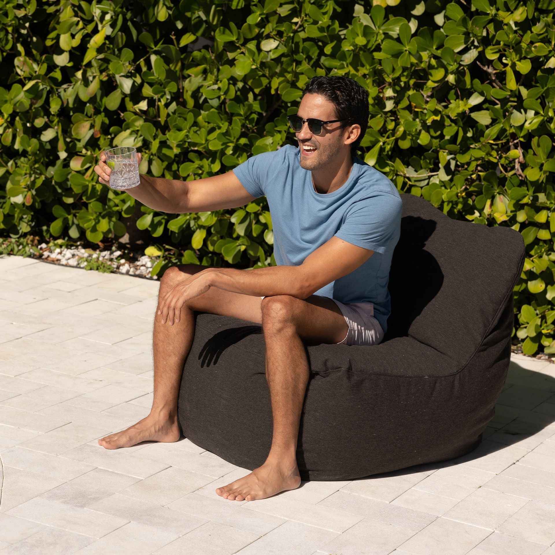 Man sitting on a brown bean bag chair outdoors, holding a drink.