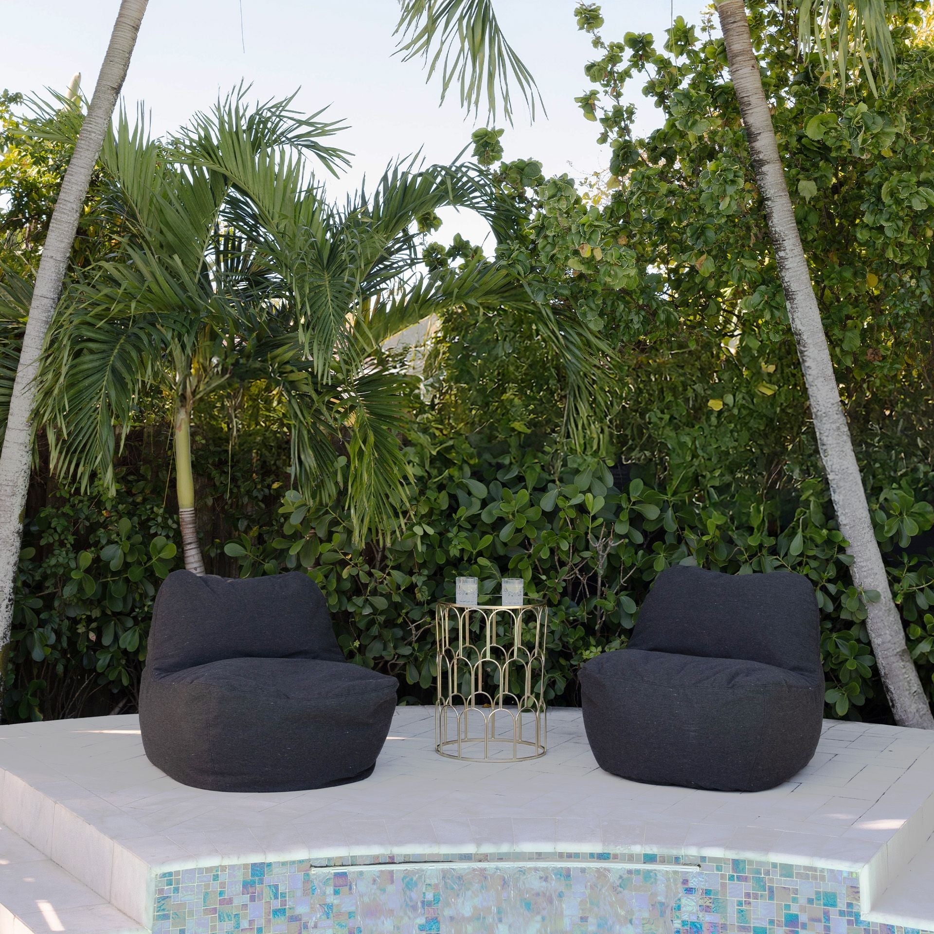 Two black bean bag chairs on a poolside deck with greenery in the background.