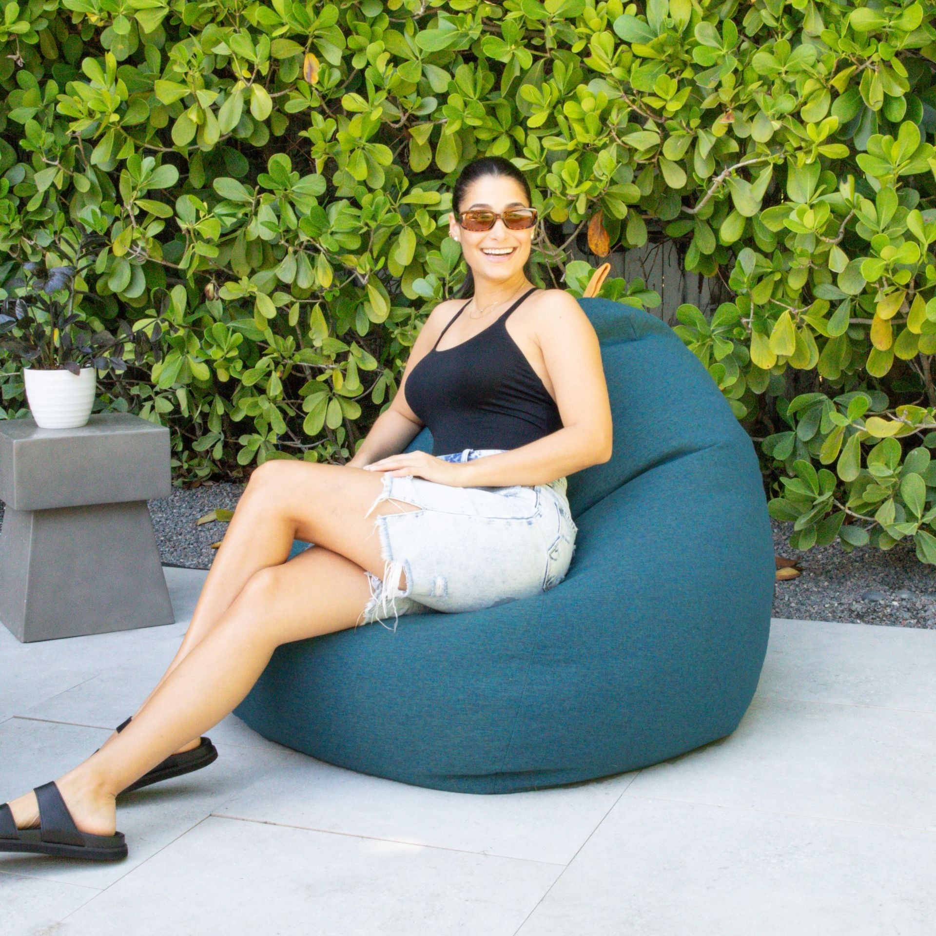 Woman sitting on a teal bean bag chair outdoors with greenery in the background