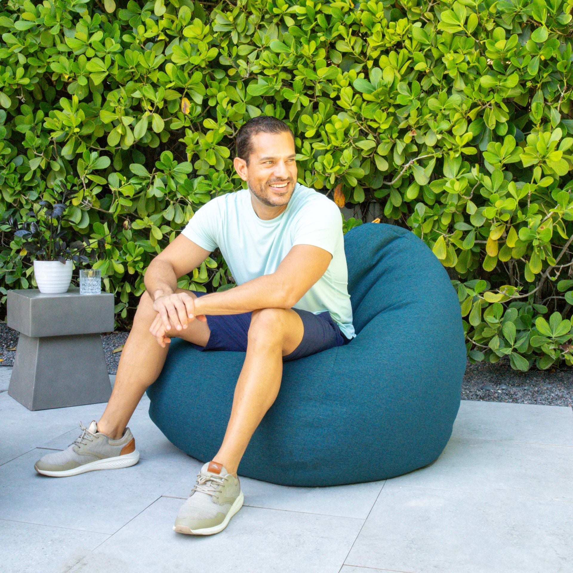 Man sitting on a teal bean bag chair outdoors with greenery in the background