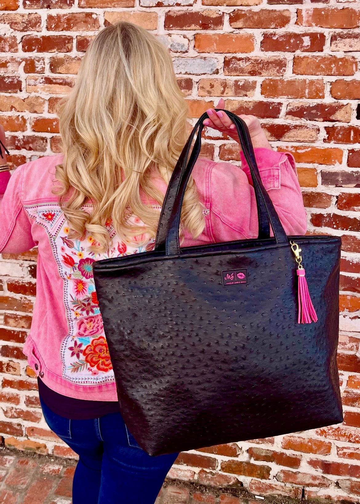 Woman holding a black textured tote bag with a pink tassel against a brick wall.
