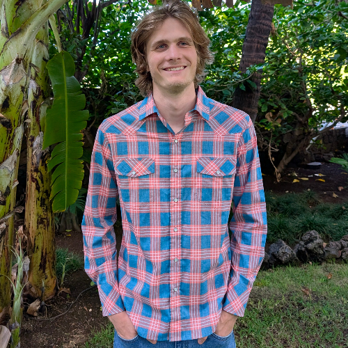 Man wearing a blue and pink plaid shirt standing outdoors with greenery in the background