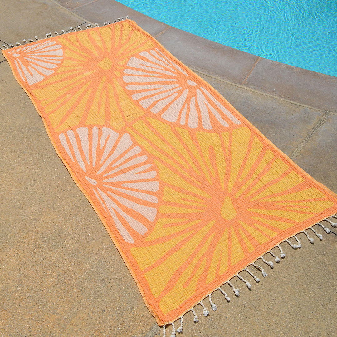 Orange and yellow patterned towel with white tassels on a stone surface near a pool.