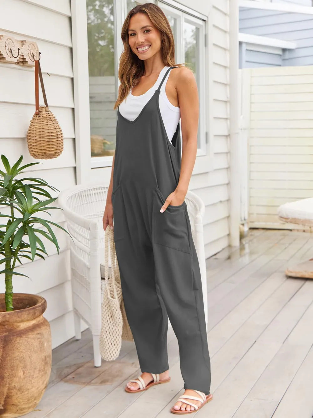 A woman standing on a patio wearing a black sleeveless jumpsuit with a white tank top underneath, holding a wicker handbag and wearing white sandals.