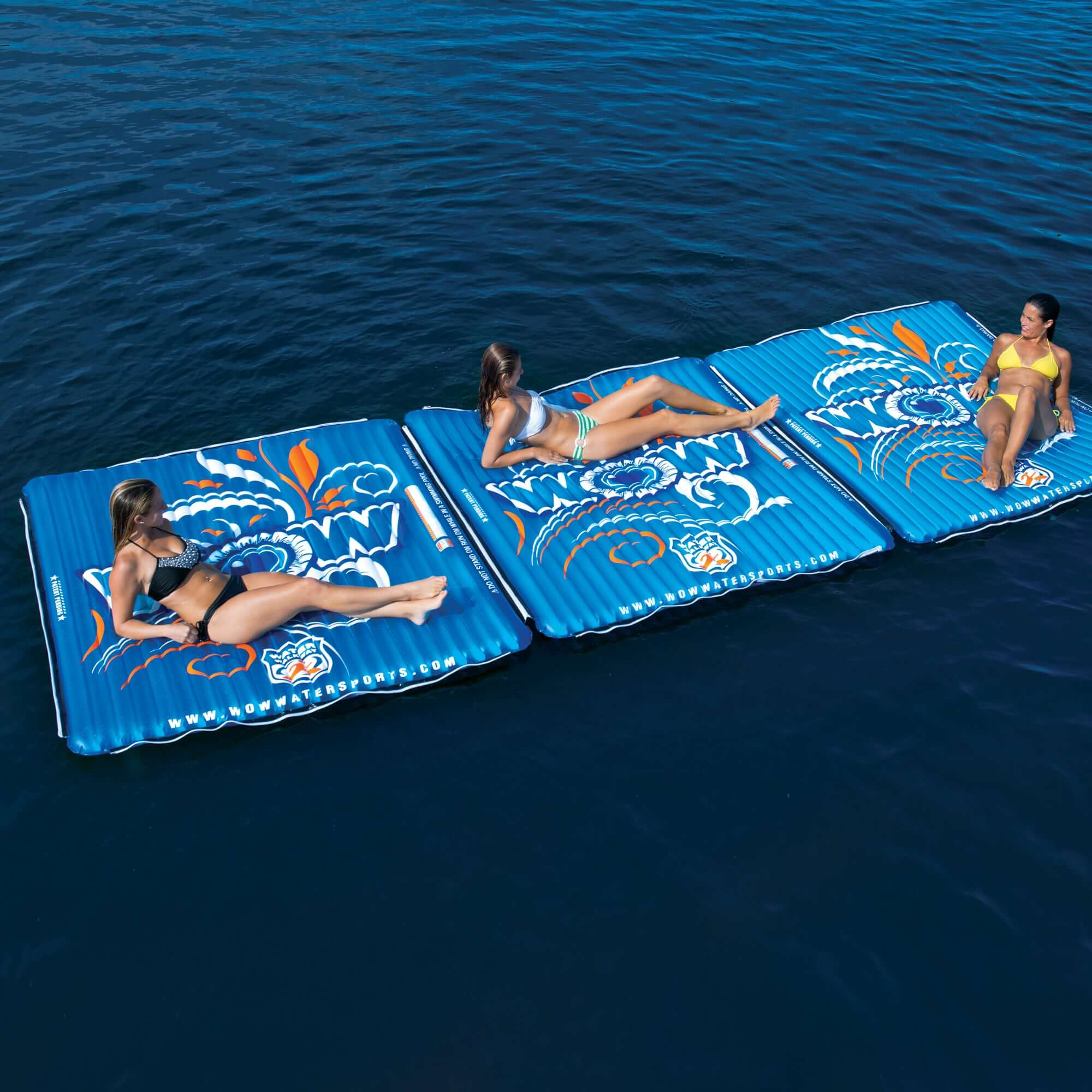 Three people lying on a blue inflatable pool mat with floral patterns in the water.