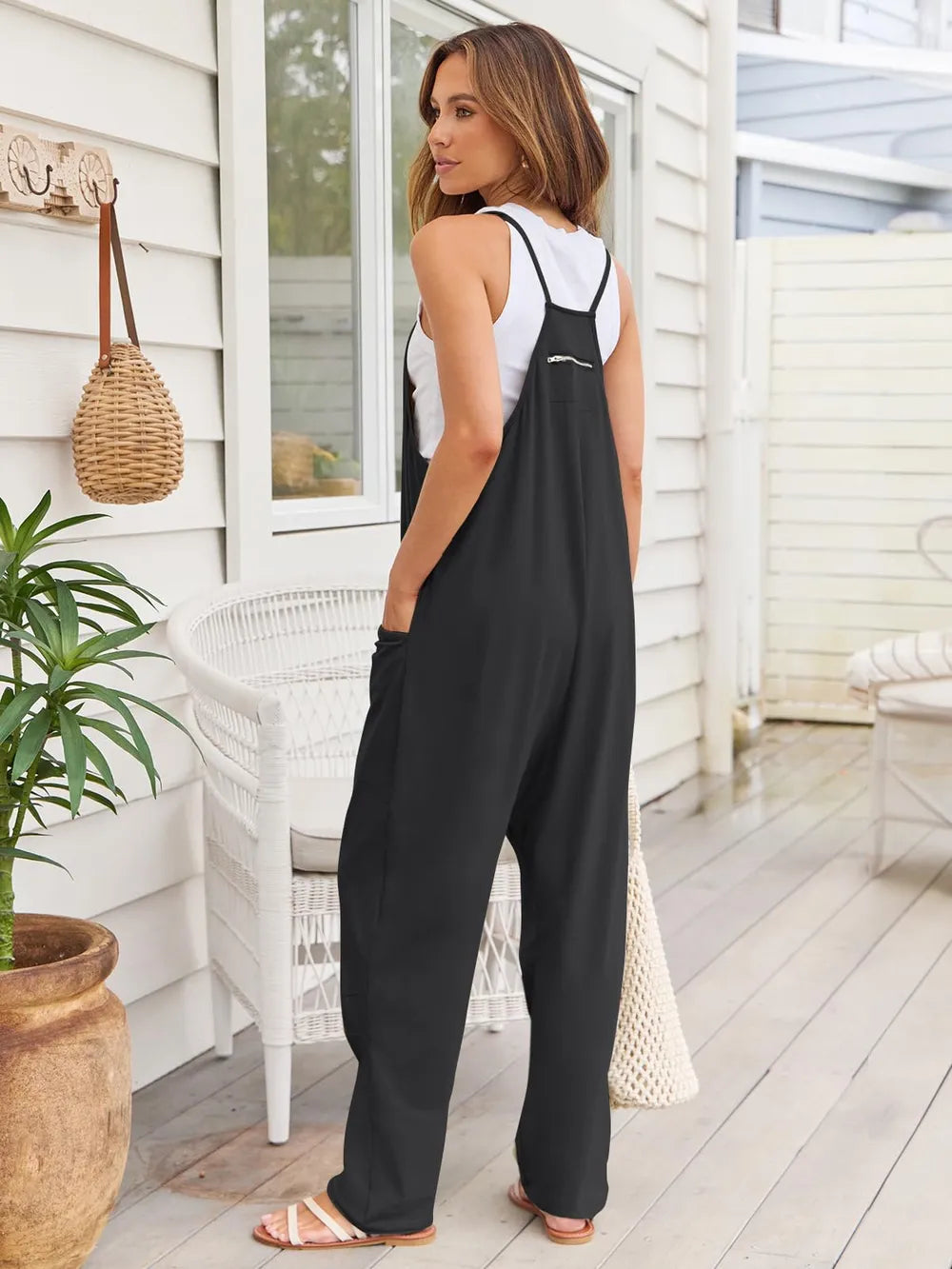 A woman standing on a patio wearing a black sleeveless jumpsuit with a white tank top underneath, holding a wicker handbag and wearing white sandals.