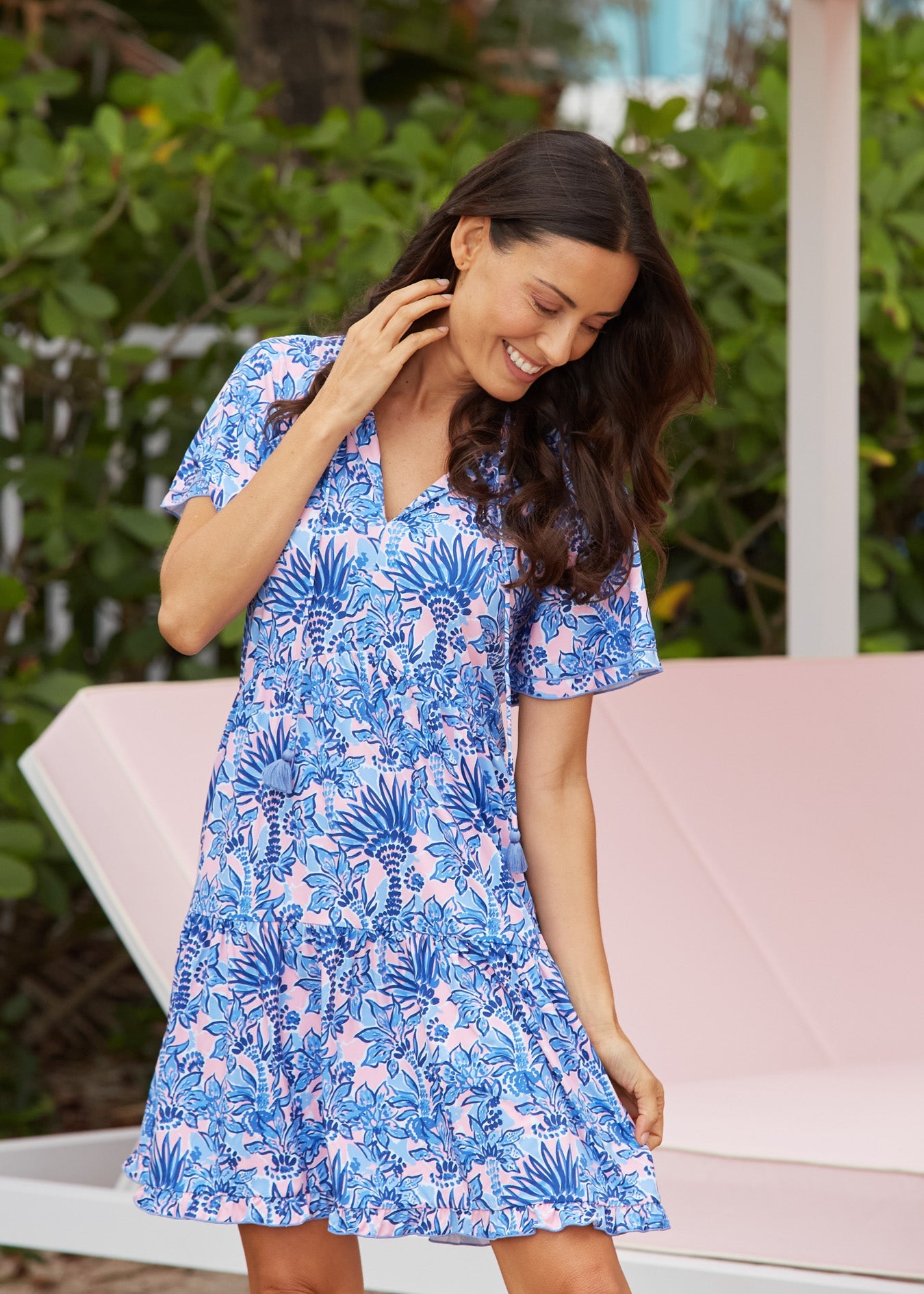 Woman wearing a blue floral dress sitting on a pink chair with greenery in the background