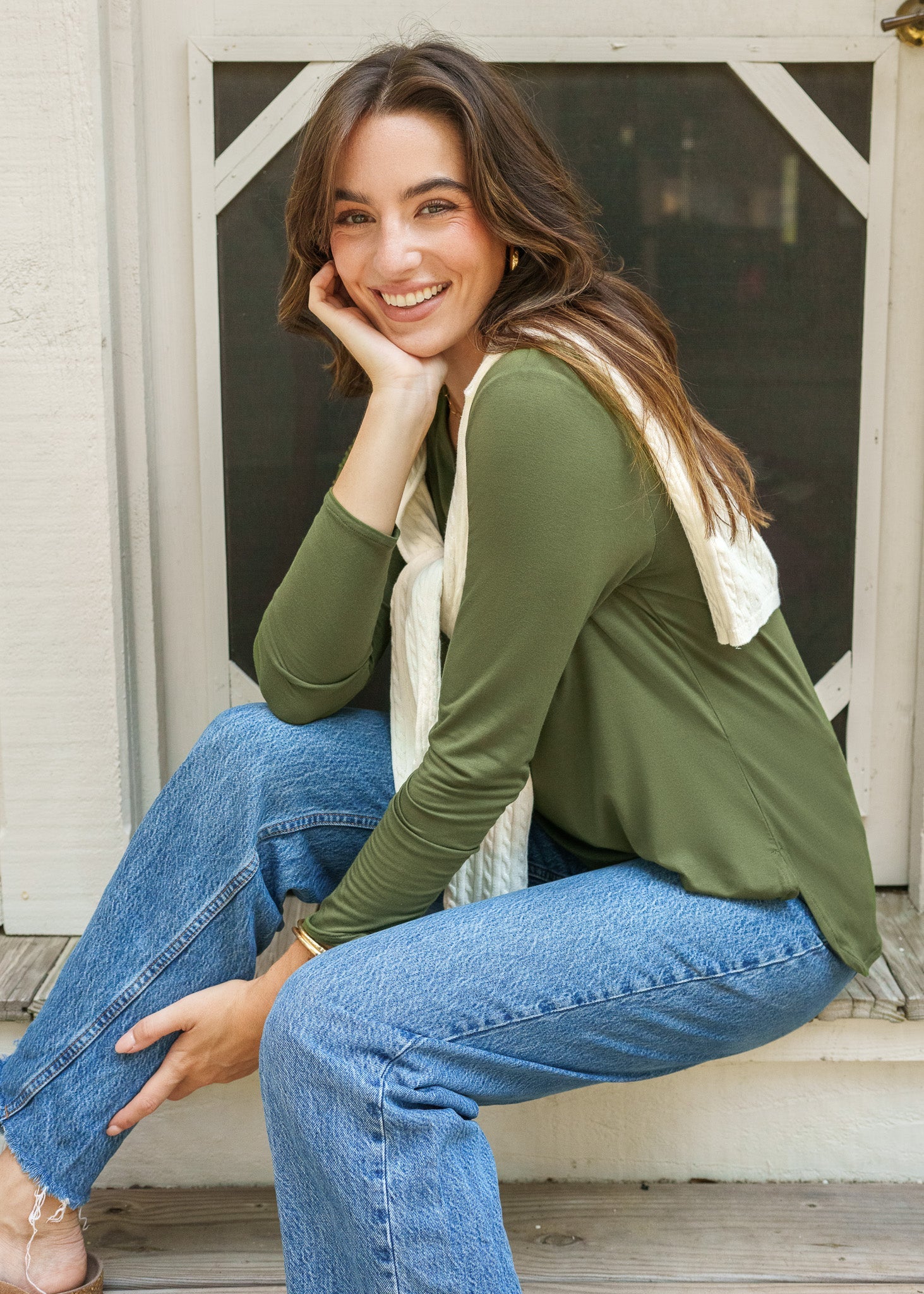 Woman sitting on a wooden step wearing a green sweater and blue jeans, smiling.