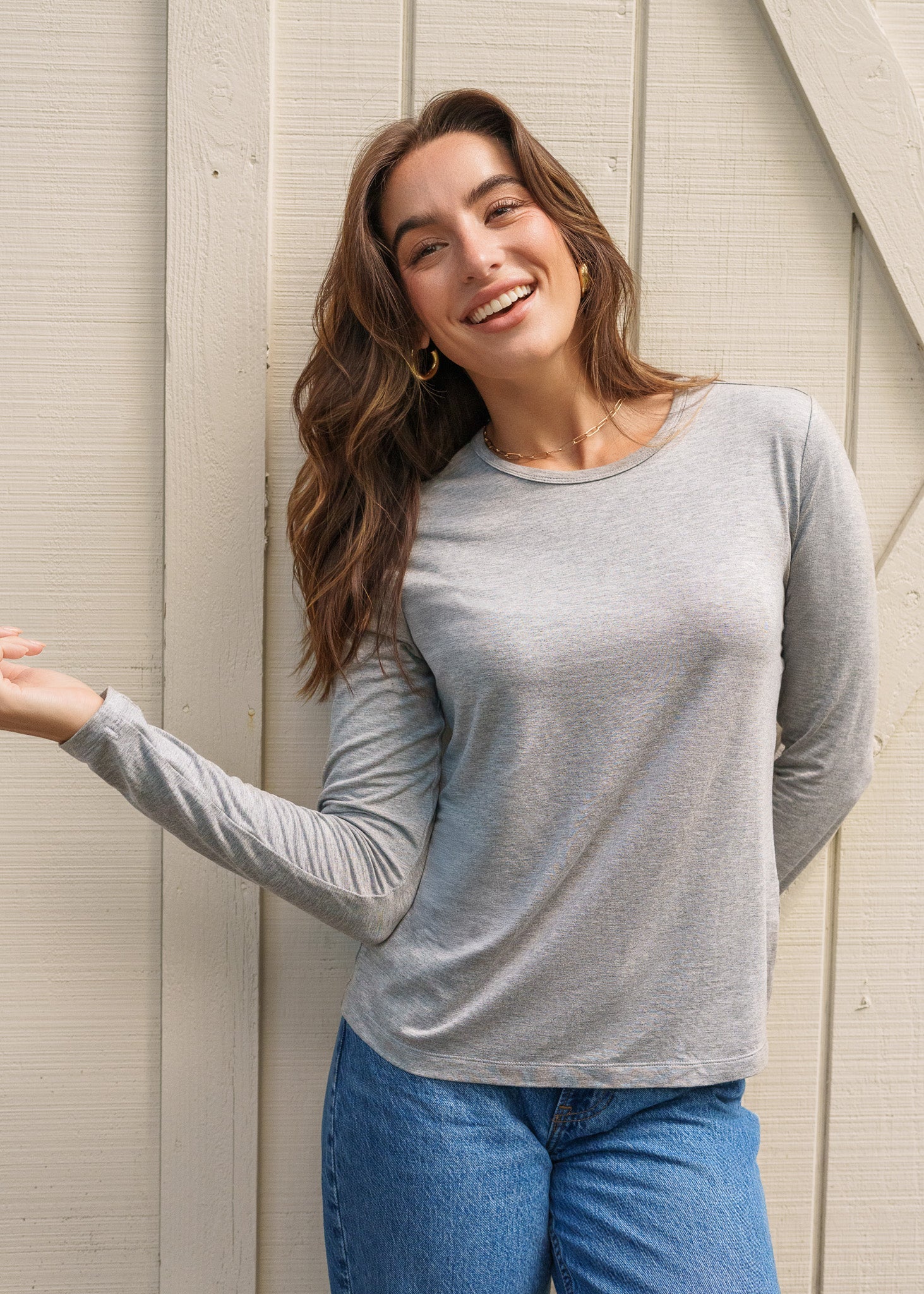 Woman wearing a gray long-sleeve shirt and blue jeans standing against a light-colored wall.