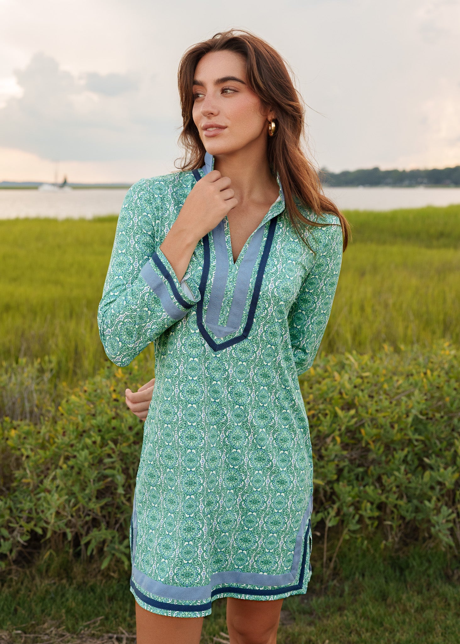 Woman wearing a green dress with blue trim standing in a grassy field with water in the background