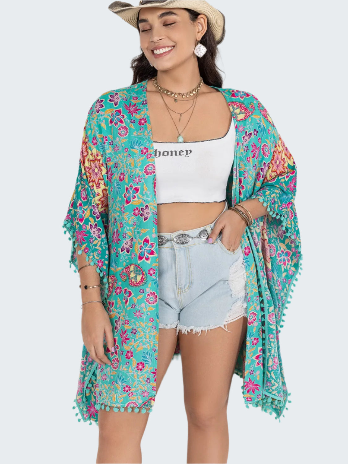 A woman wearing a multicolored floral print open front cover-up with tassel detailing, paired with a white top and blue denim shorts.