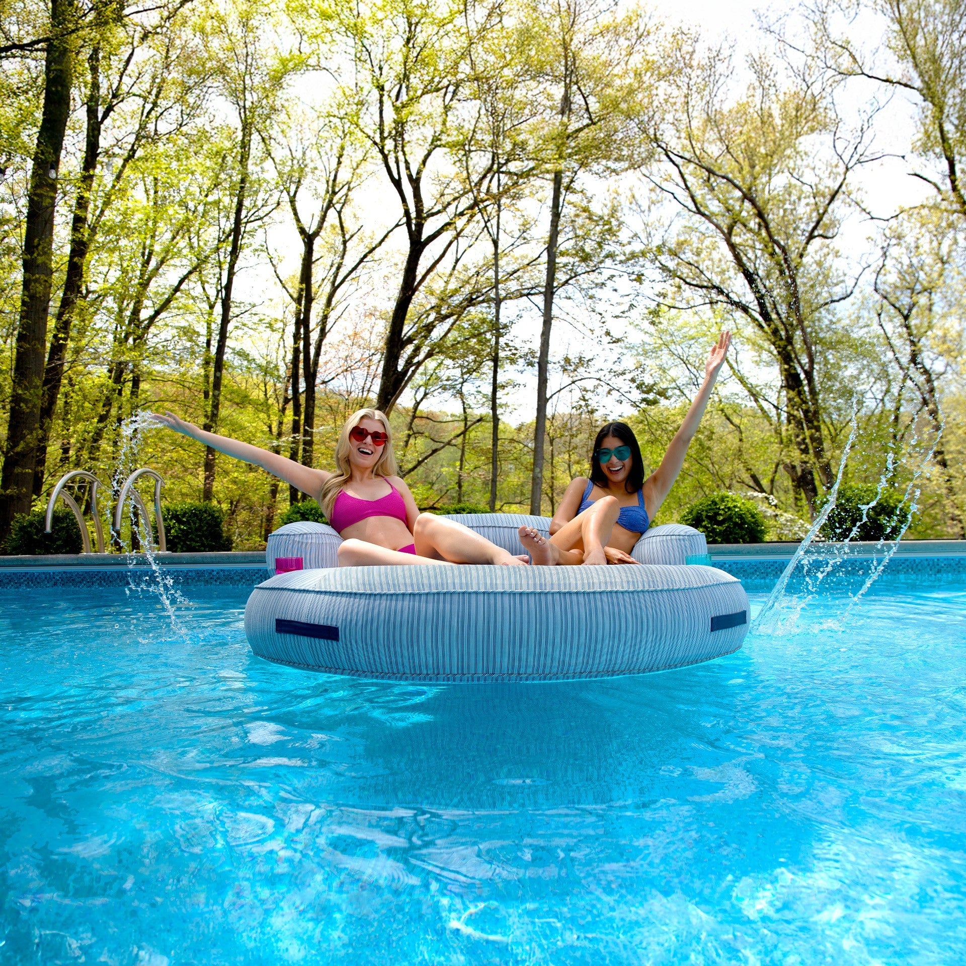 Two women relaxing on an inflatable pool float in a pool surrounded by trees.