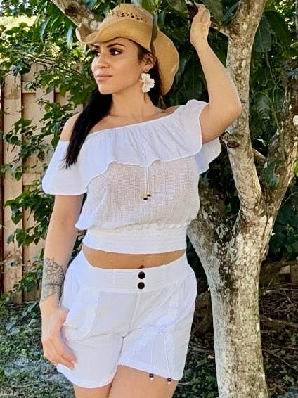 Woman in a white outfit with a straw hat standing outdoors near trees.
