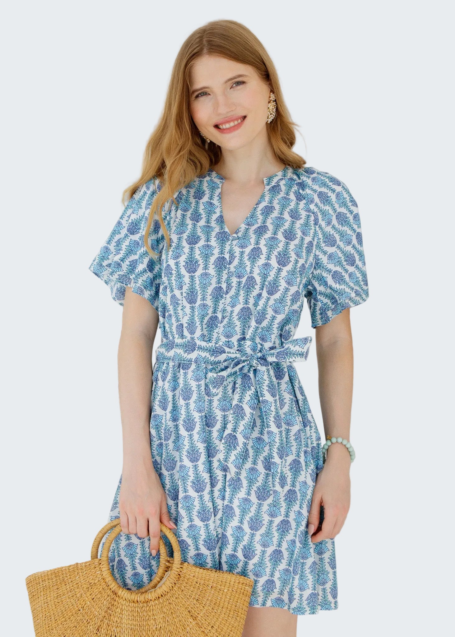 Woman wearing a blue patterned dress holding a straw bag against a white wall.