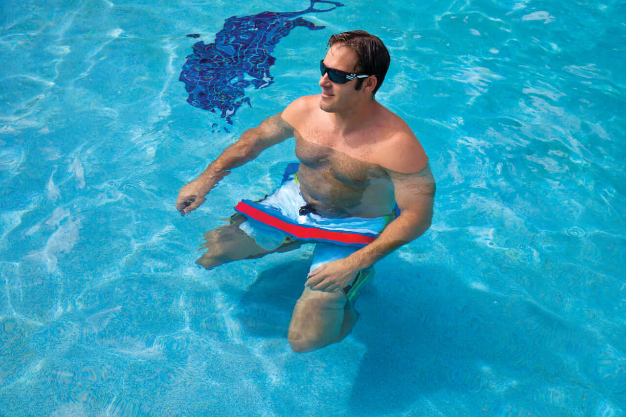 Man in a pool wearing sunglasses and swim shorts with a red stripe.