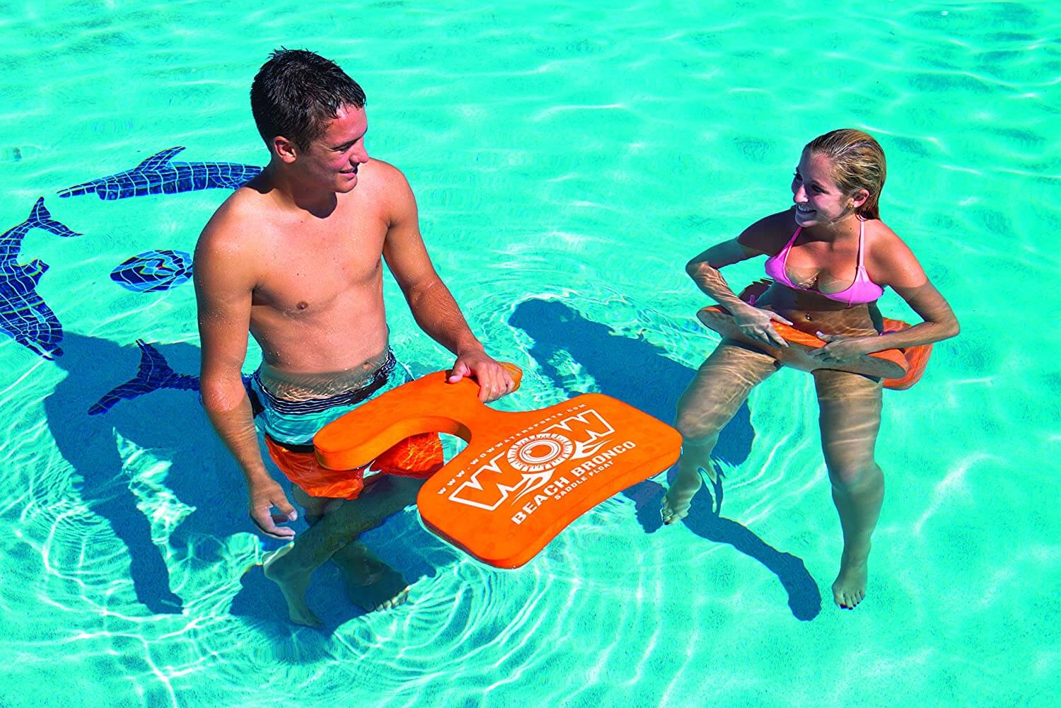 Two people holding orange bodyboards in a pool with shark-shaped floats in the background.